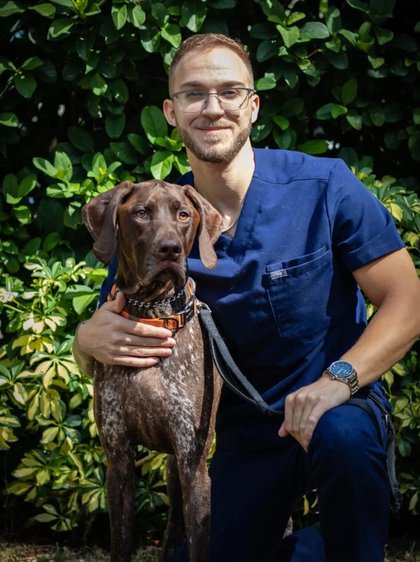 Man in blue scrubs kneeling with a brown dog outdoors